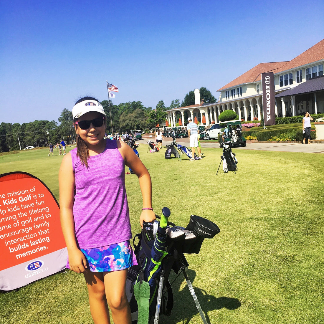 A smiling young girl golfer in a purple tank top and visor stands with her golf bag at a  Golf tournament, with a large white clubhouse and golf carts in the background in a sunny day..